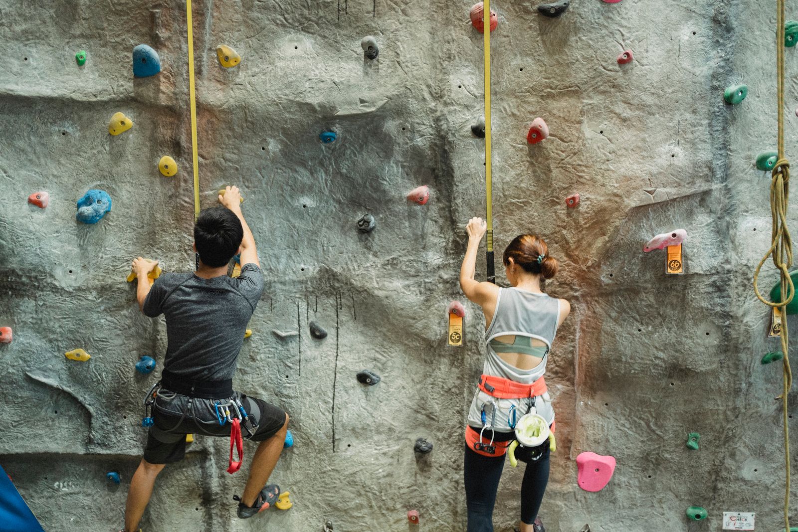 A climber in a harness on a tall wall with a belayer holding the rope below